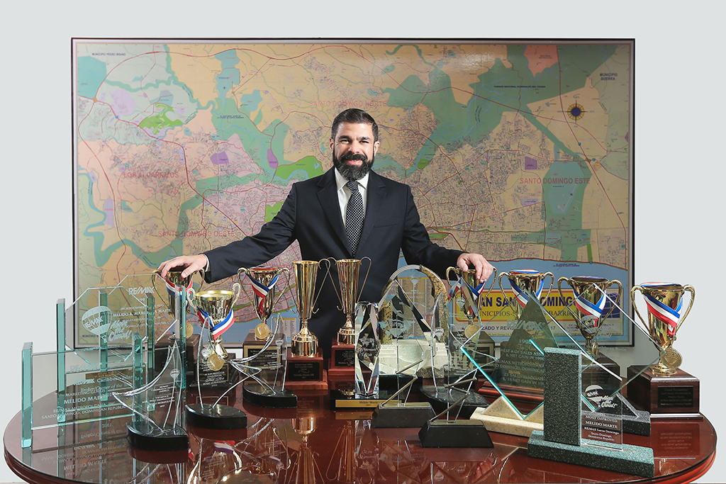 Melido Marte detras de una mesa que tiene todos los trofeos y galardones que ha recibido como agente inmobiliario